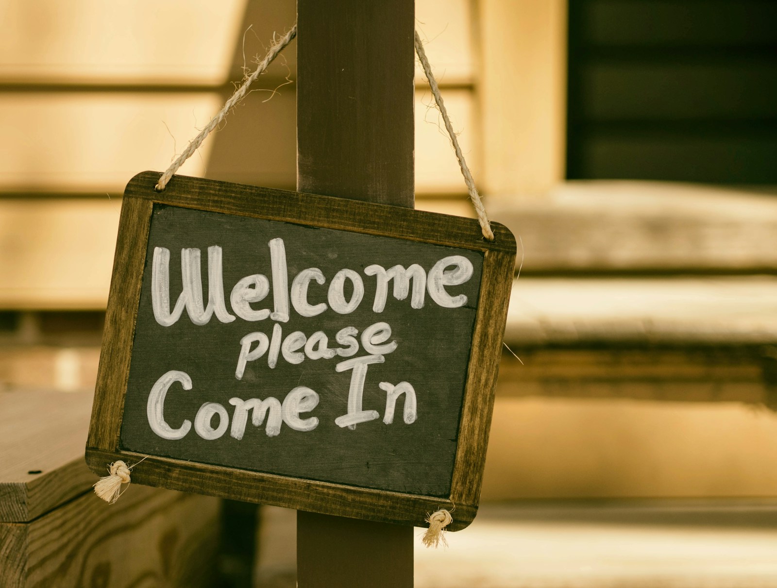 A wooden 'Welcome, please come in' chalkboard sign hanging from a porch post